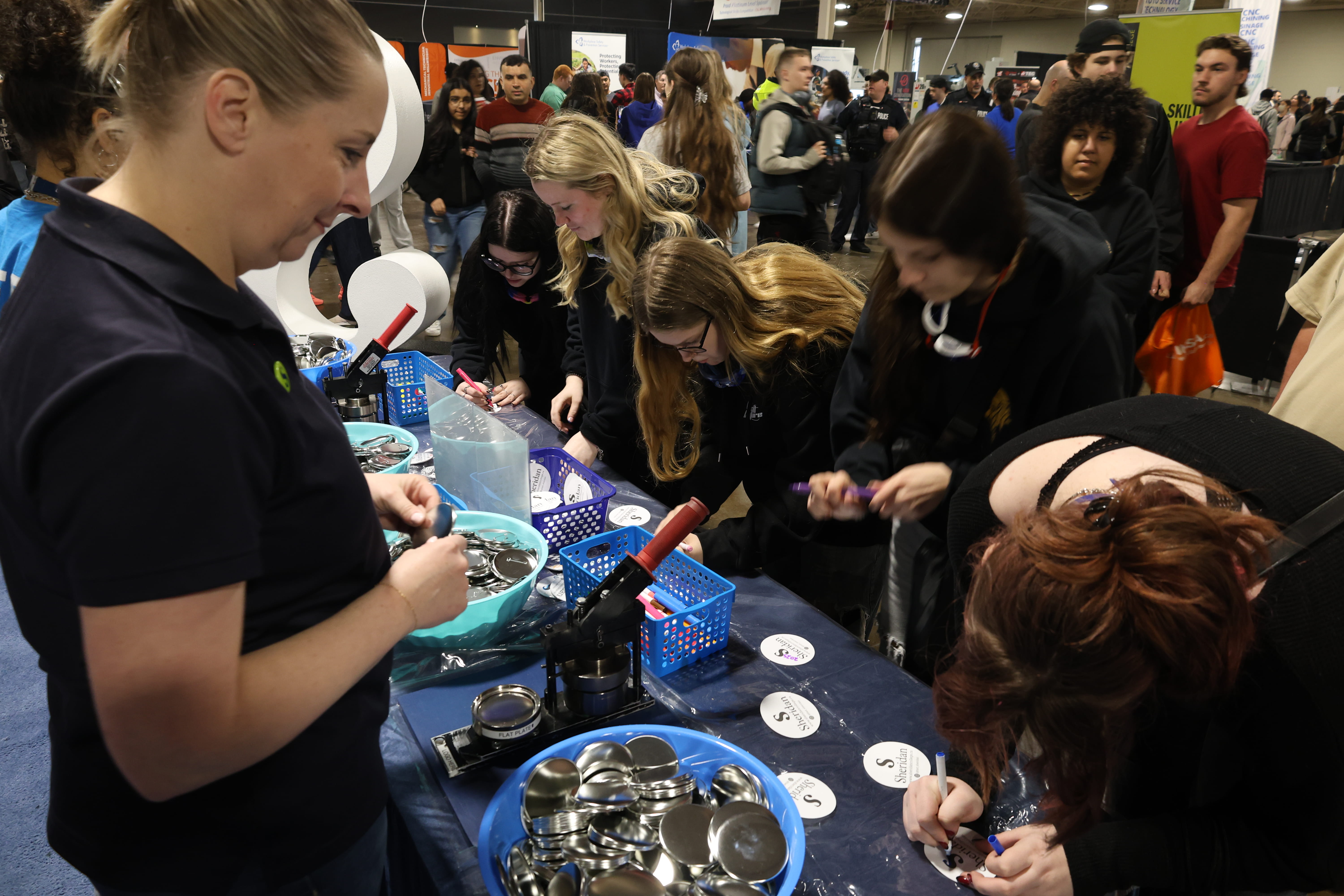 A Sheridan staff member stands behind a desk giving away pens, pins and lanyards to visitors to Sheridan's Skills Ontario booth