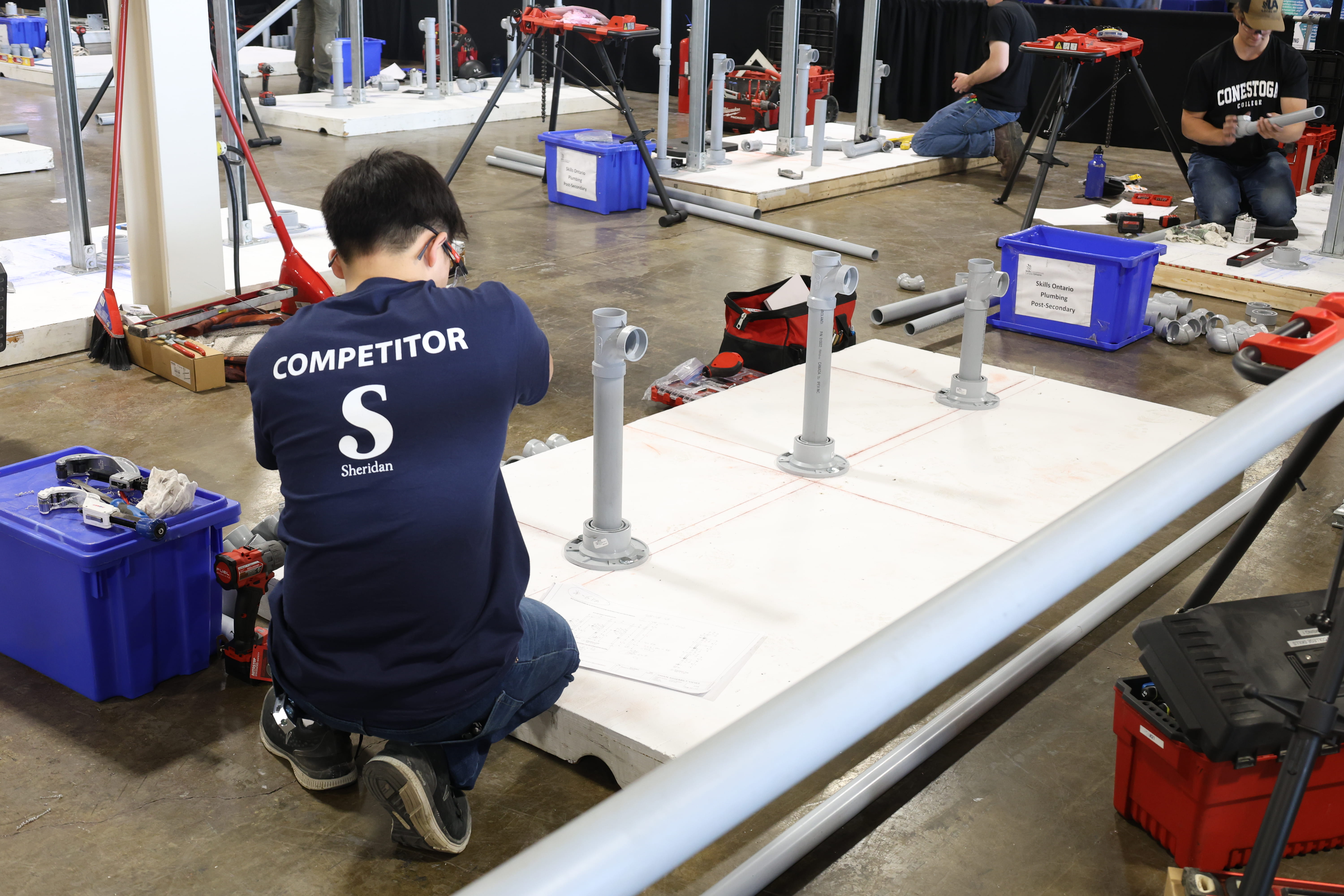 A Sheridan student crouches on the floor while working on a plumbing challenge