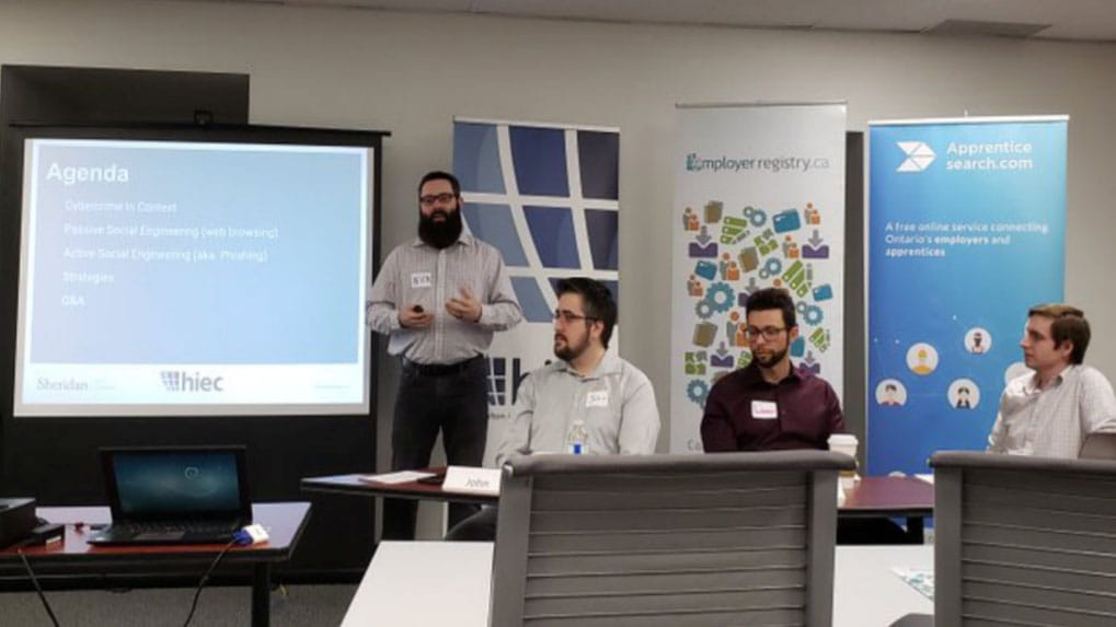Nick Johnston standing next to a large display screen with three people seated to his right.