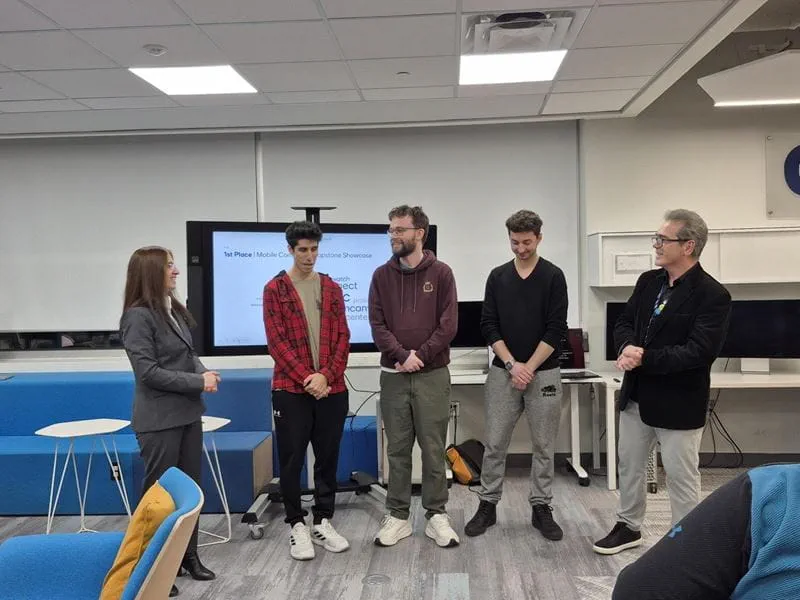 DistantSync team members Omar Al-Dulaimi, Jacob Collins and Elias Alissandratos stand at the front of the room in front of a large TV to receive congratulations from professors Shalini Singh Jaspal and Magdin Stoica