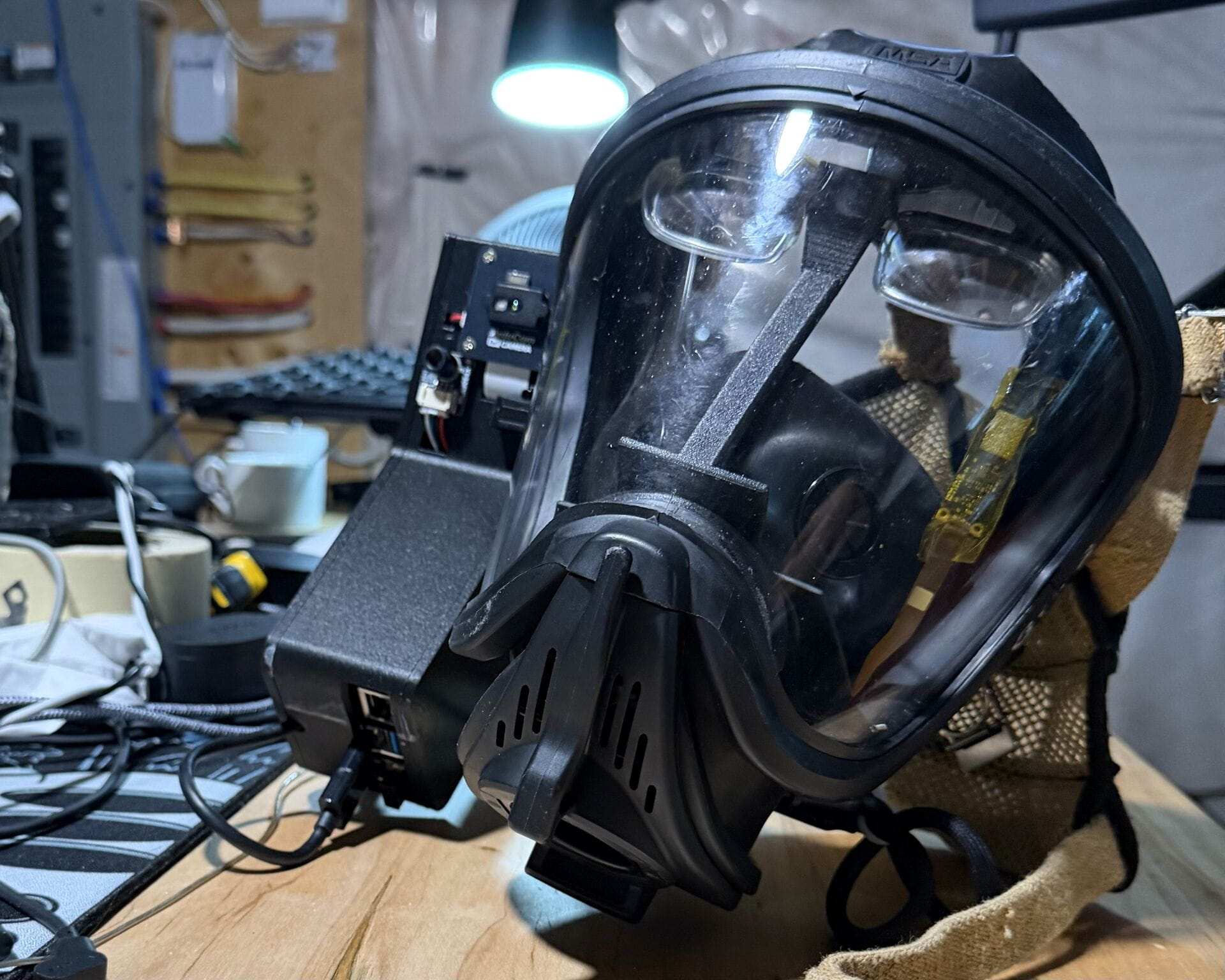 Close-up of a full-face respirator mask resting on a workbench beside electronic equipment and cables during prototype development.