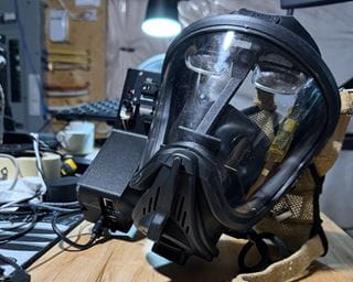 Close-up of a full-face respirator mask resting on a workbench beside electronic equipment and cables during prototype development.