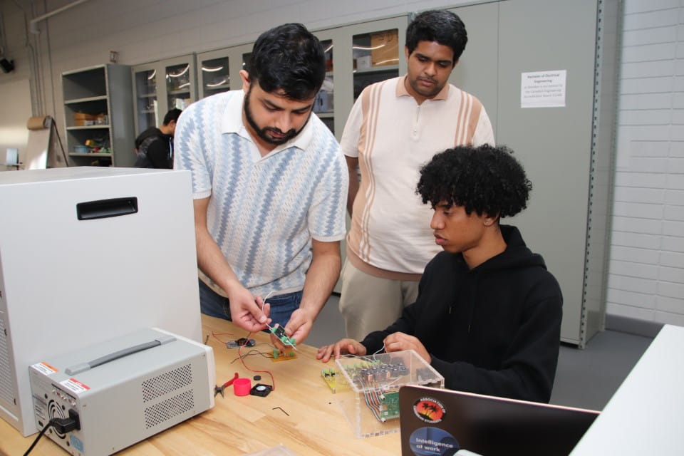 Three students stand at a desk working on their levitation-based conveyor system.
