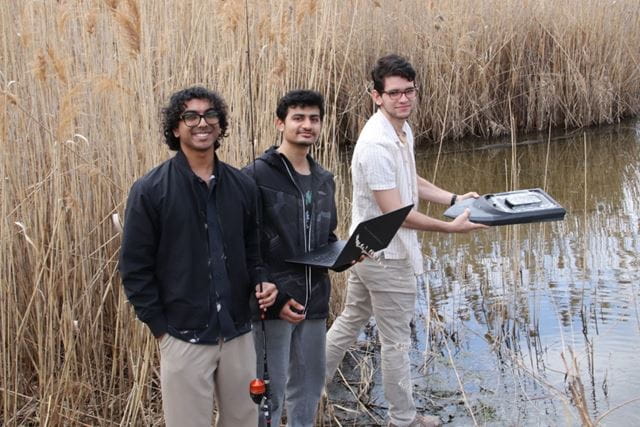 Three students pose with their boat while standing next to the pond on Sheridan's Davis Campus.