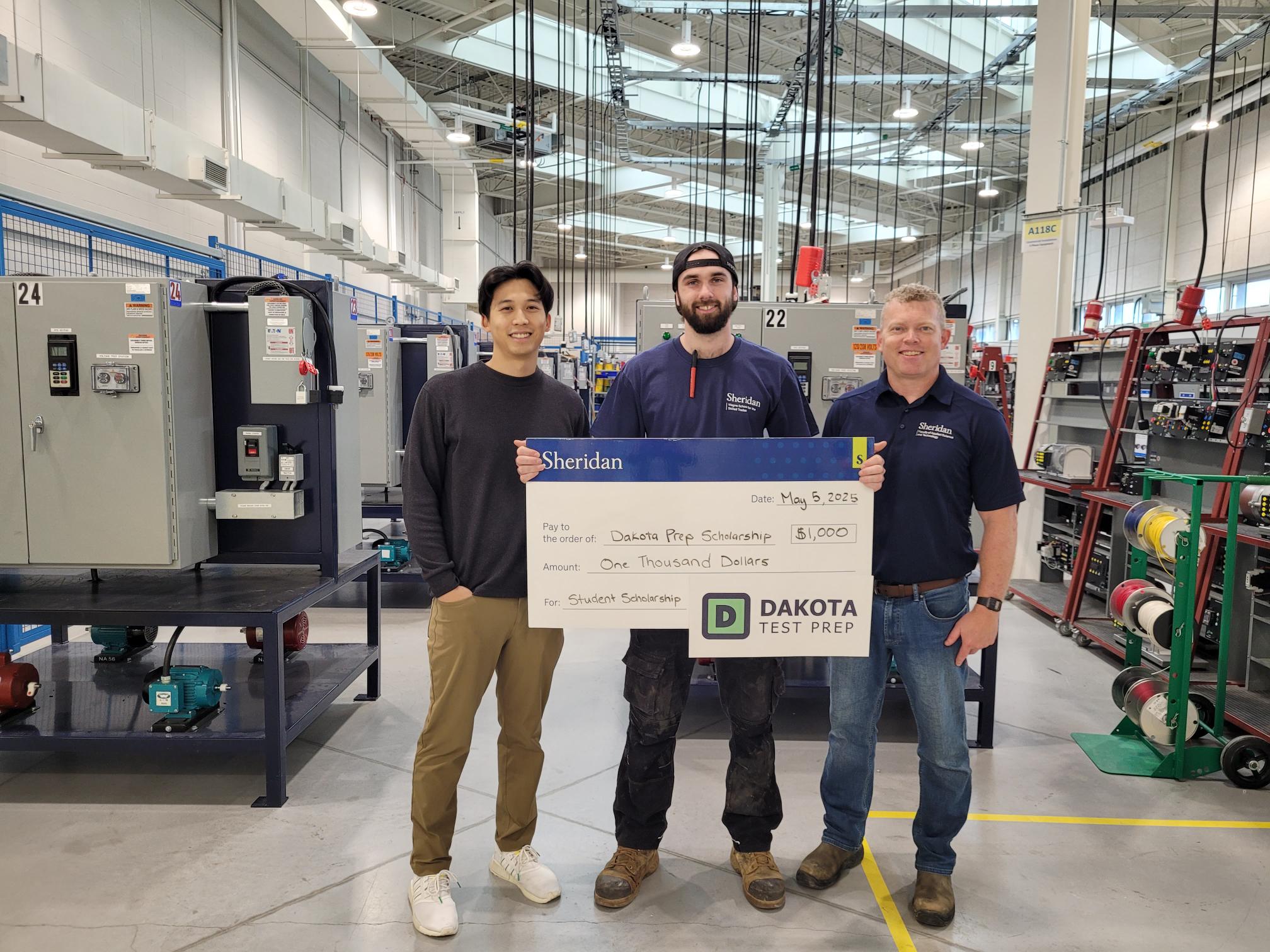 Sheridan Construction and Maintenance Electrician apprentice Keaton Snow holds an oversized cheque as he stands in one of Sheridan's lab spaces between Dakota Prep founder Alex Au and Sheridan program coordinator Dan Hammond