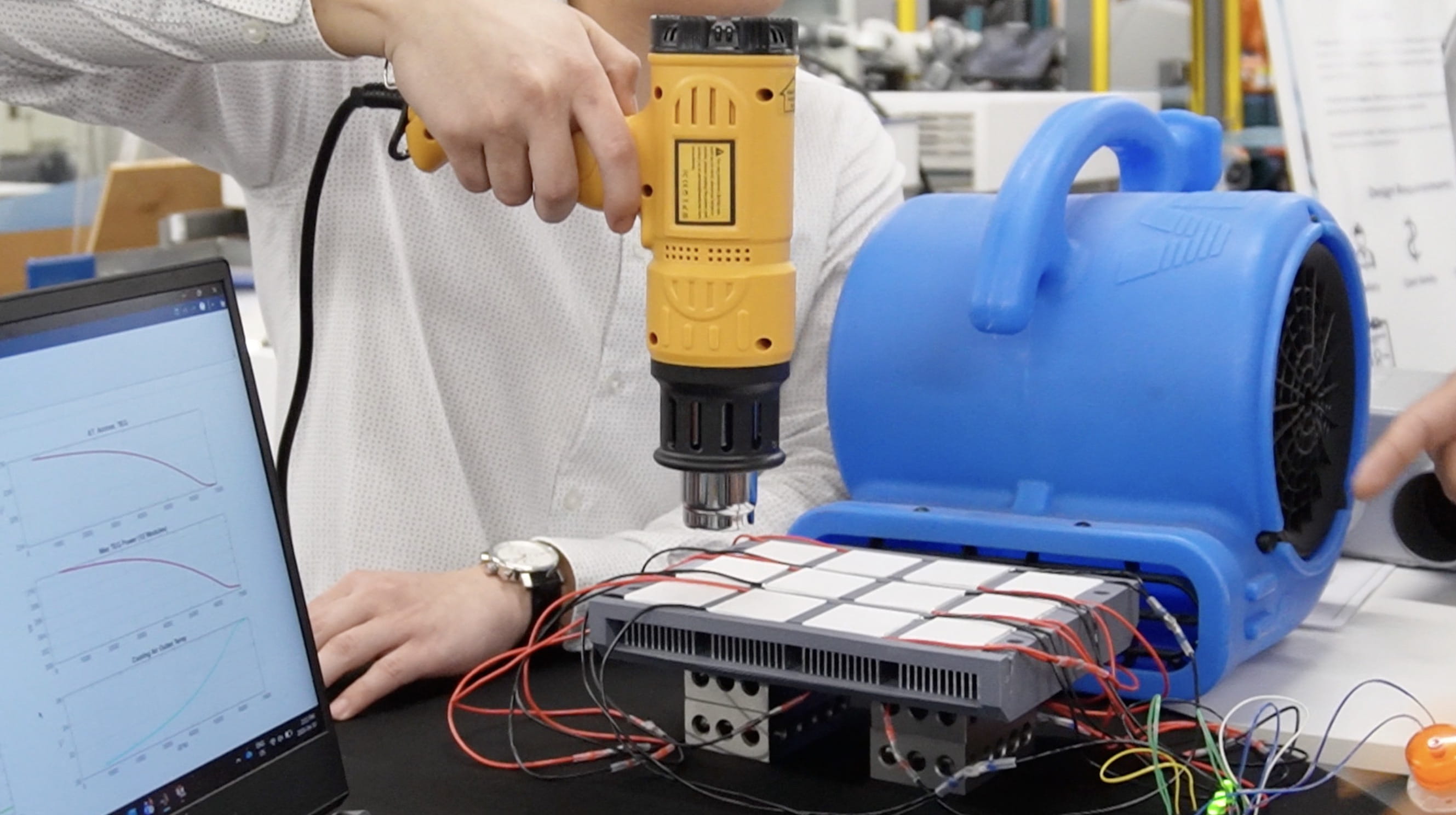 A heat gun is pointed at heat syncs and modules to simulate the temperature of a car, with a fan pointed towards the setup to simulate driving conditions.