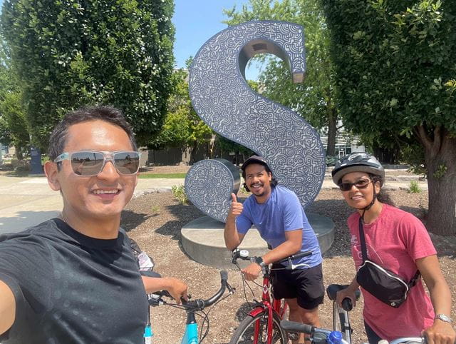 Three cyclists stand together in front of the large blue  S logo at Davis campus.