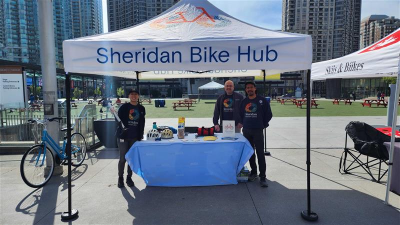 Three people stand at a table under the Sheridan bike hub tent