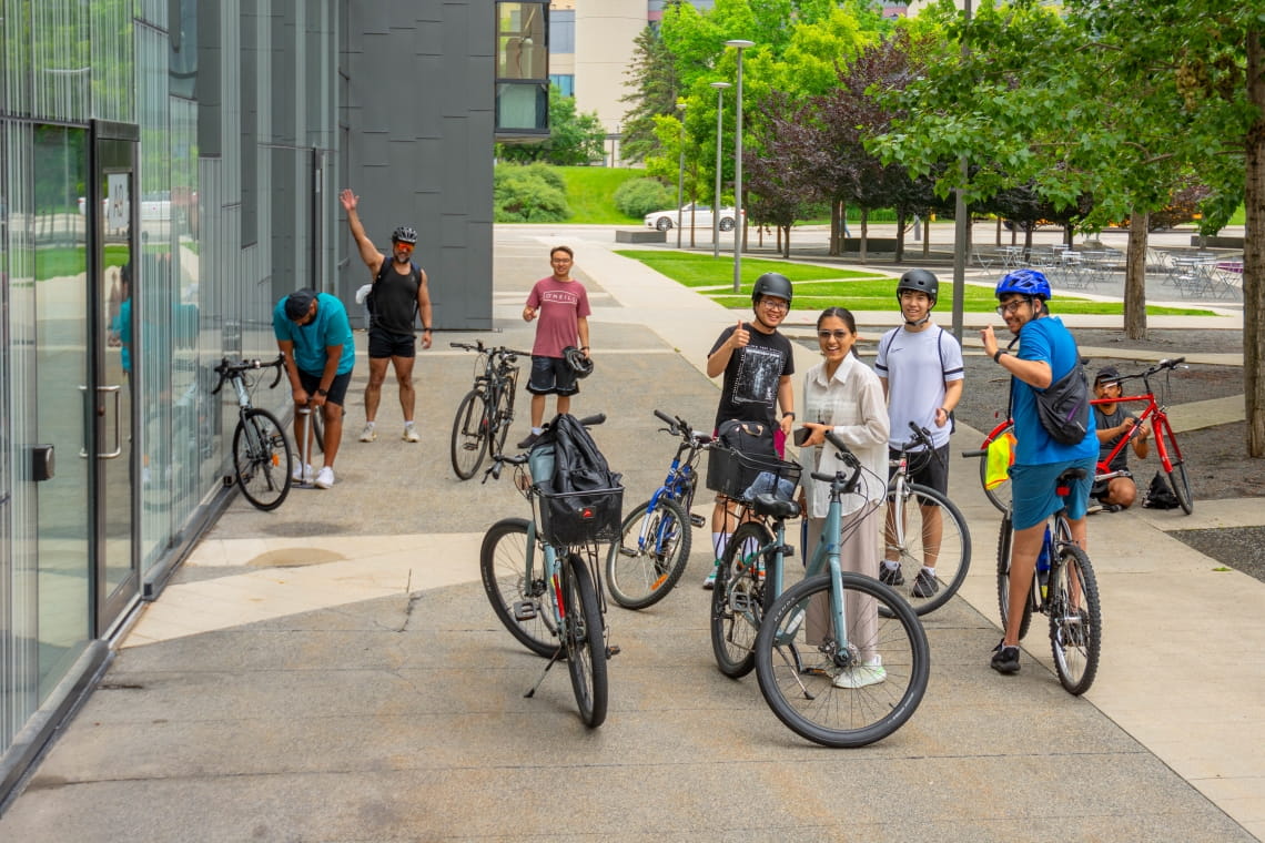 A group of people stand with their bikes on the sidewalk on campus