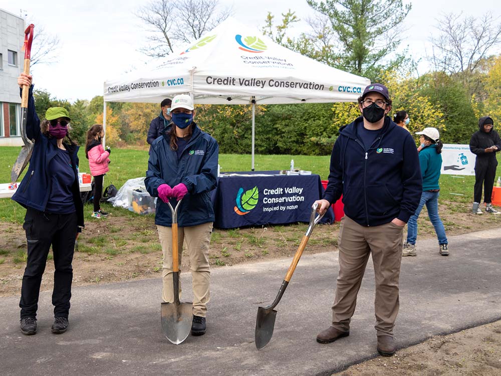 A group of three volunteers with shovels in front of the Credit Valley Conservation tent