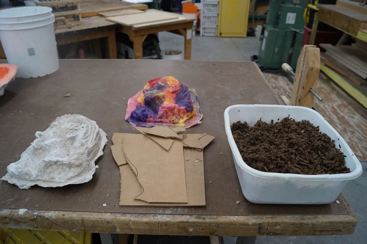 A bin of wet paper scraps sits on a table next to scraps of cardboard and student art projects.