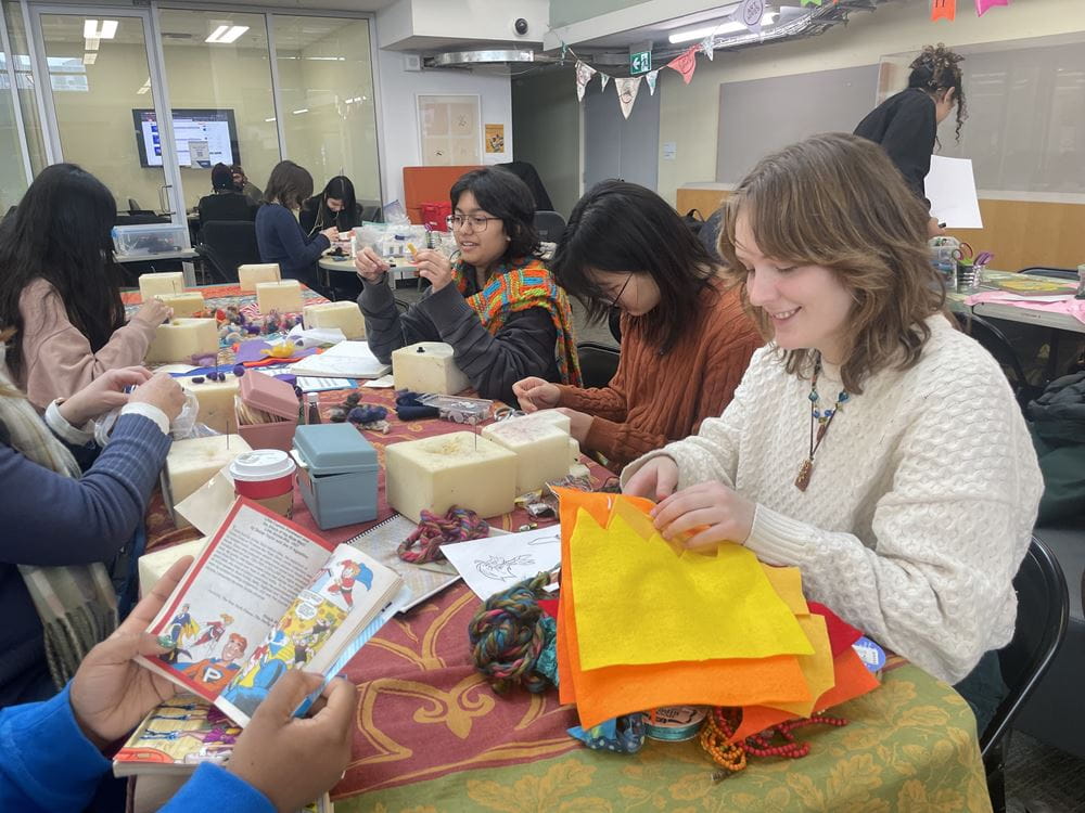 Several students sit at a table which is covered with various arts and crafts supplies.