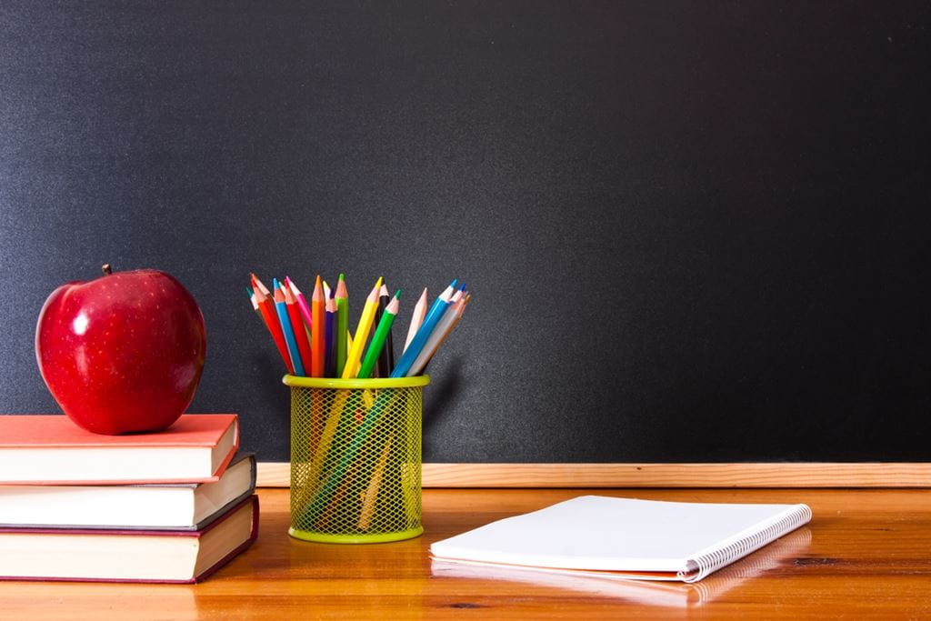 An illustration of a teacher's desk in a classroom. There are pencil crayons, papers and an apple on the desk, and a chalkboard in the background.