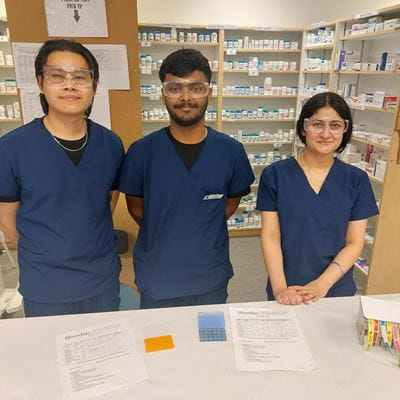 Three Pharmacy Technician students stand behind a counter in Sheridan's pharmacy lab, with pharmaceutical products pictured behind them on shelves