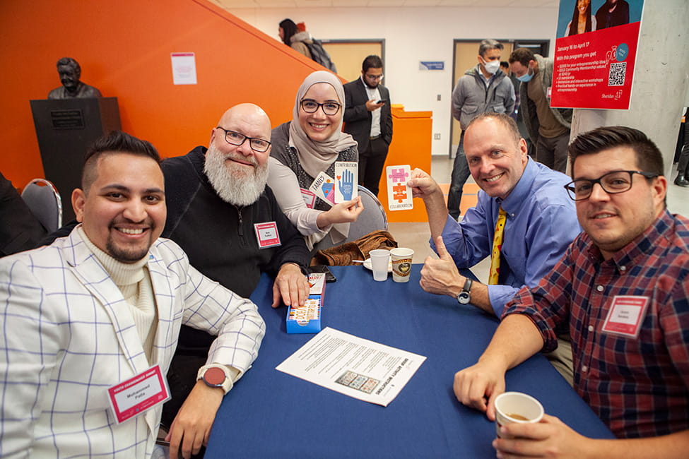 People seated around a table and smiling. Two are holding up three cards that read 'FAMILY', 'CONTRIBUTION', and 'COLLABORATION'