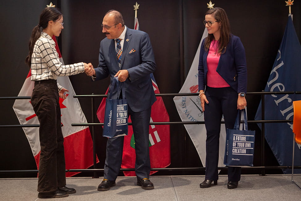 Two people to the left shaking hands. One person standing off to the right observing them.