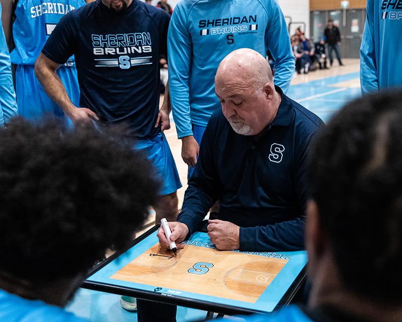 Sheridan men's basketball coach Jim Flack draws up a play while his players watch