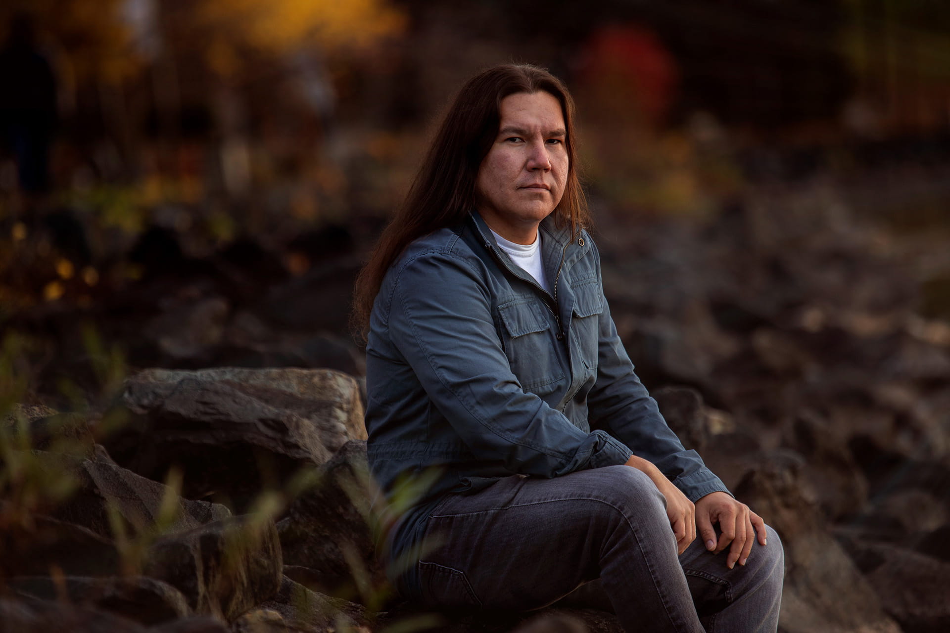 Celebrated Anishinaabe author Waubgeshig Rice poses for a photo while seated outside on a large boulder