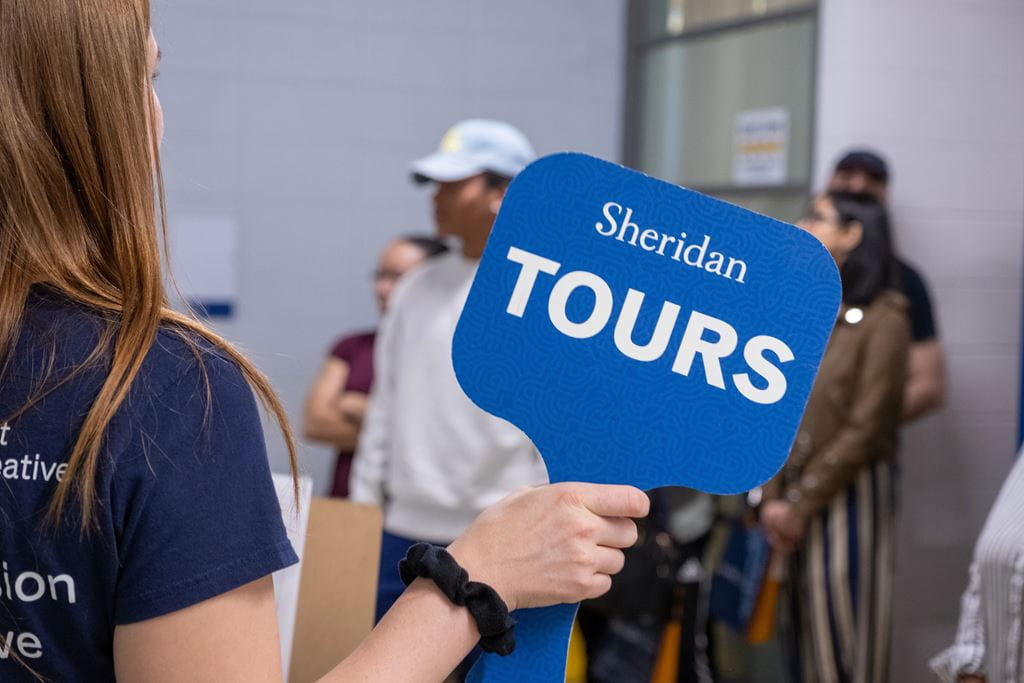 A Sheridan student ambassador holds up a "Sheridan Tours" sign during an on-campus tour.