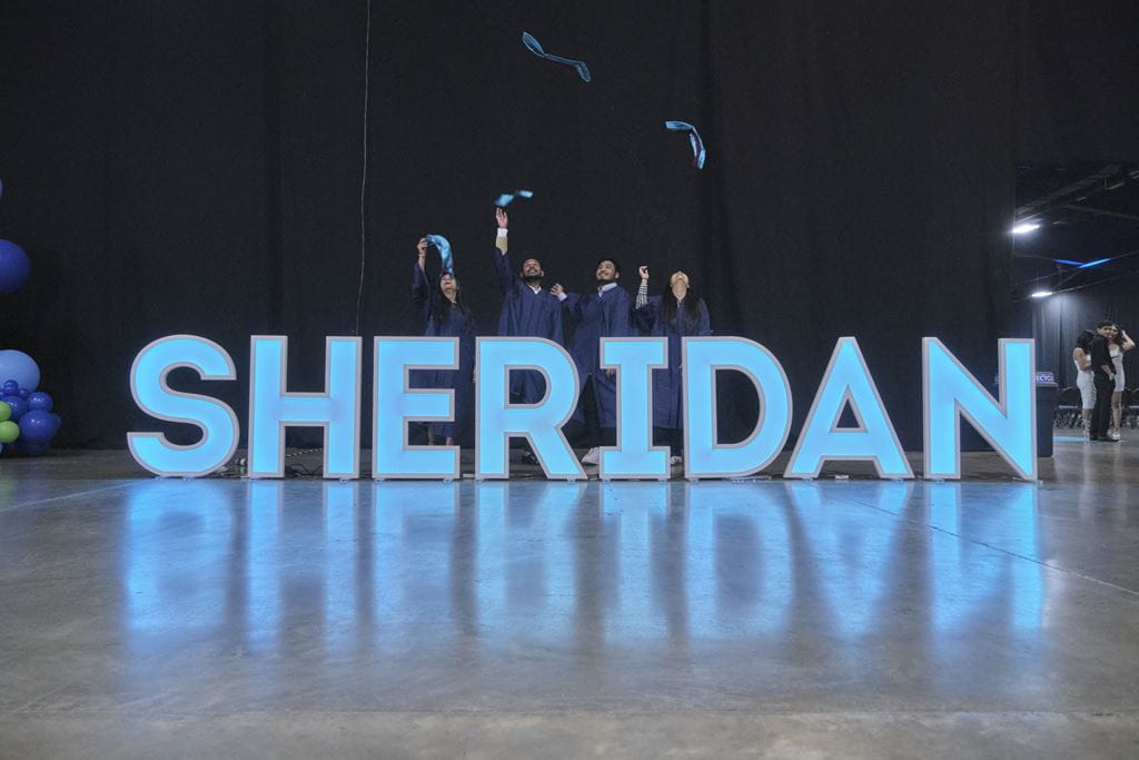 Graduates stand behind a sign that spells out the name SHERIDAN in all capital letters