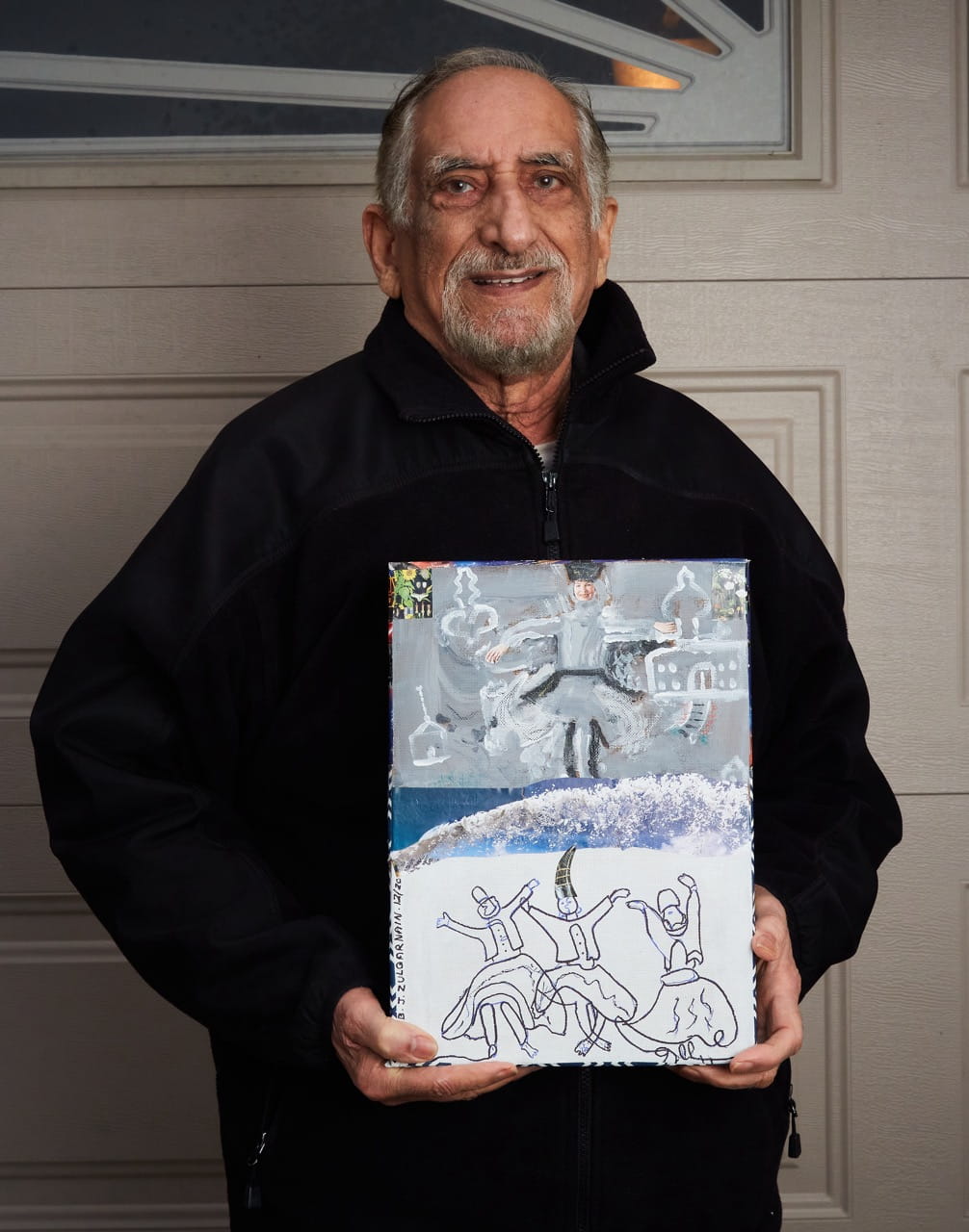 An older adult smiles while holding up a piece of artwork he created.