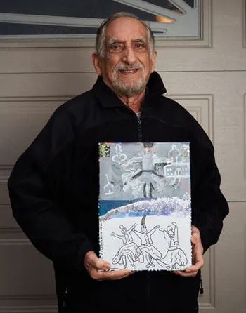 An older adult smiles while holding up a piece of artwork he created.