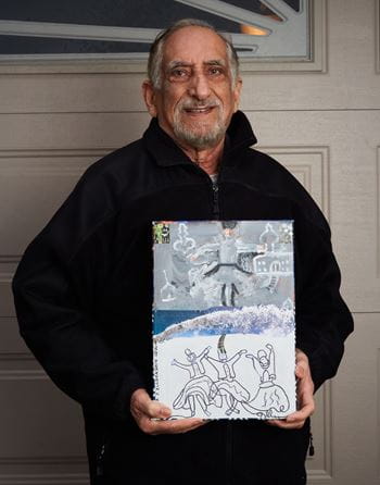 An older adult smiles while holding up a piece of artwork he created.