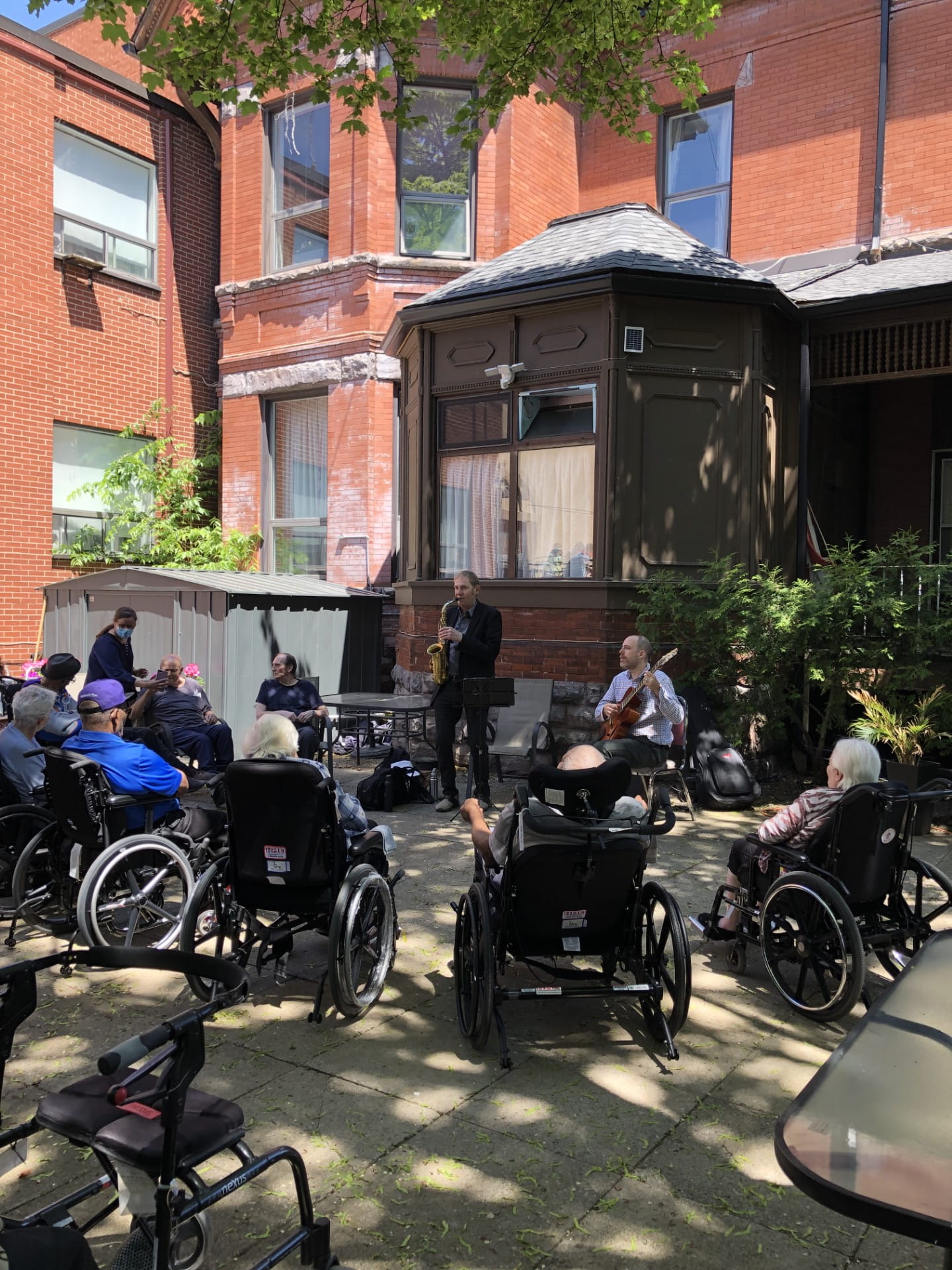 A group of older adults sit outside in a semicircle watching a saxaphonist and guitarist perform.