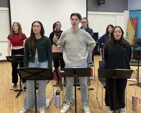 A group of six students facing the camera sing as part of their rehearsal warmup
