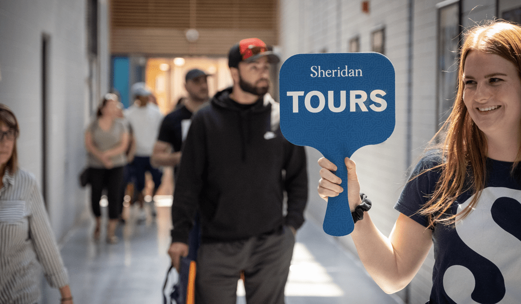A woman smiles while holding a sign that reads "Sheridan Tours" as a group of Open House attendees follow behind
