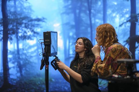 Two people stand on a production set looking at the display of a camera