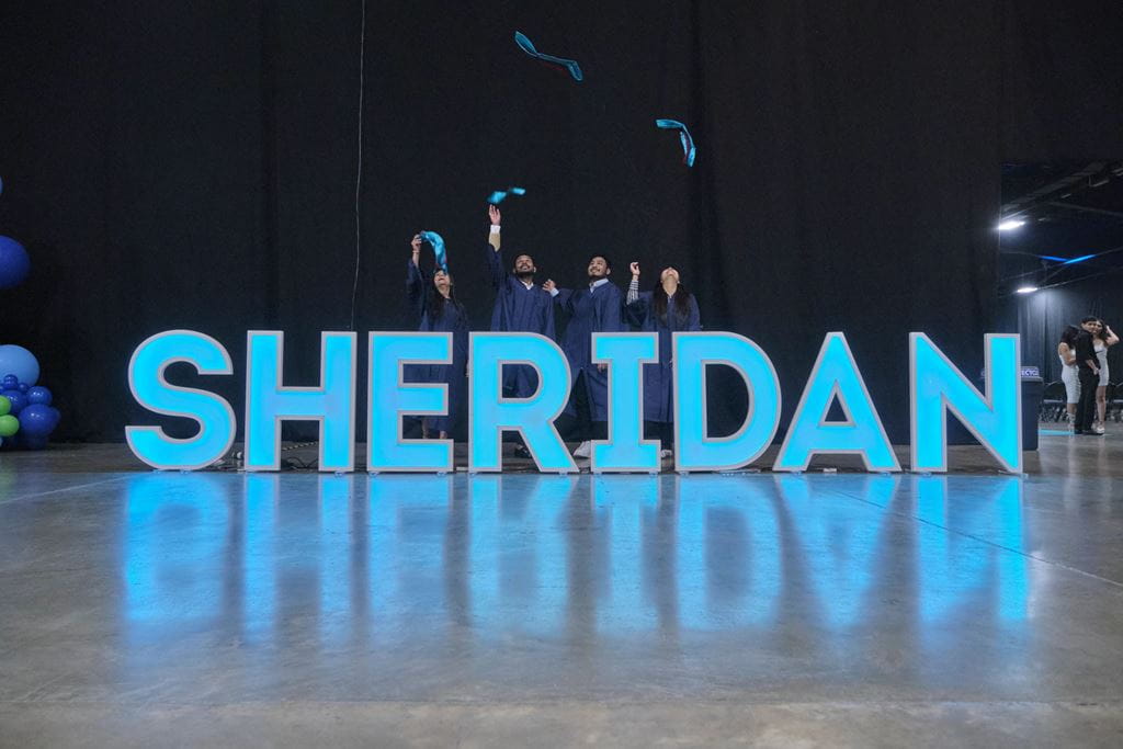 Graduates throw their caps in the air while standing behind giant letters that spell out the name SHERIDAN