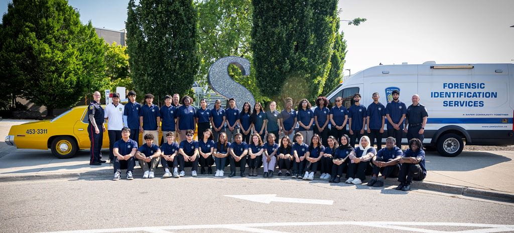 A group of 35 students wearing dark blue shirts, part of the Youth in Policing Initiative, stand with staff members in front of the large Sheridan S, a police van and a yellow car.