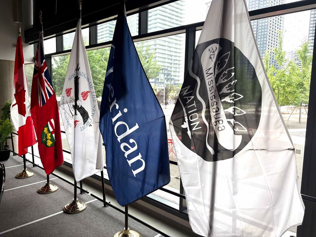 Five flags on flag poles are displayed in front of a large window at Sheridan's Hazel McCallion campus.