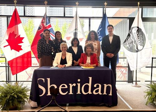A group of seven people sit and stand behind a table with the lettering Sheridan, as two people sign a memorandum of understanding document.