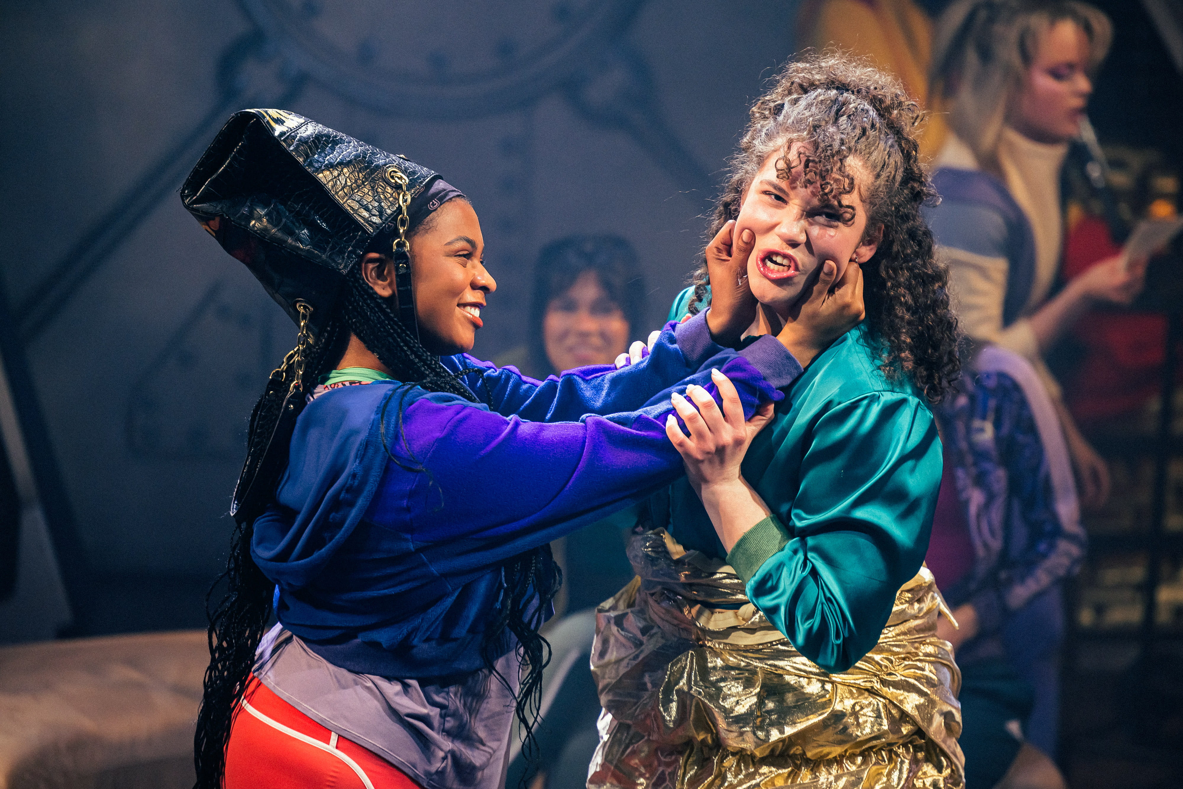 A smiling actor in costume on stage grabbing the face of a second actor, who is grimacing.