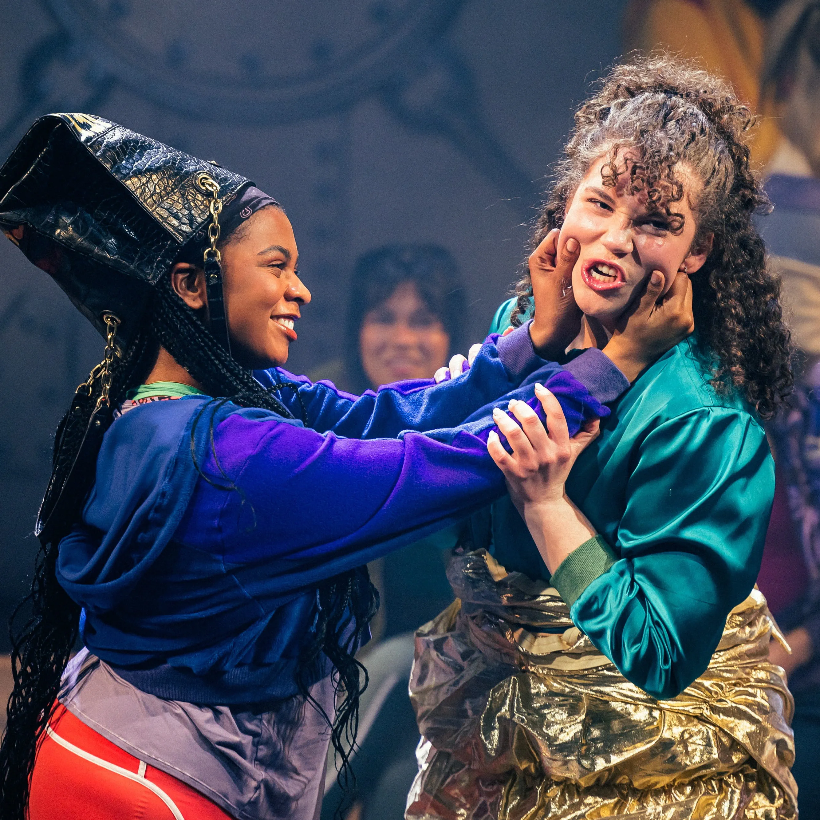 A smiling actor in costume on stage grabbing the face of a second actor, who is grimacing.