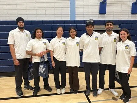 Seven students wearing white shirts stand in front of the bleachers in a gym.