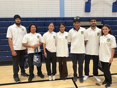 Seven students wearing white shirts stand in front of the bleachers in a gym.