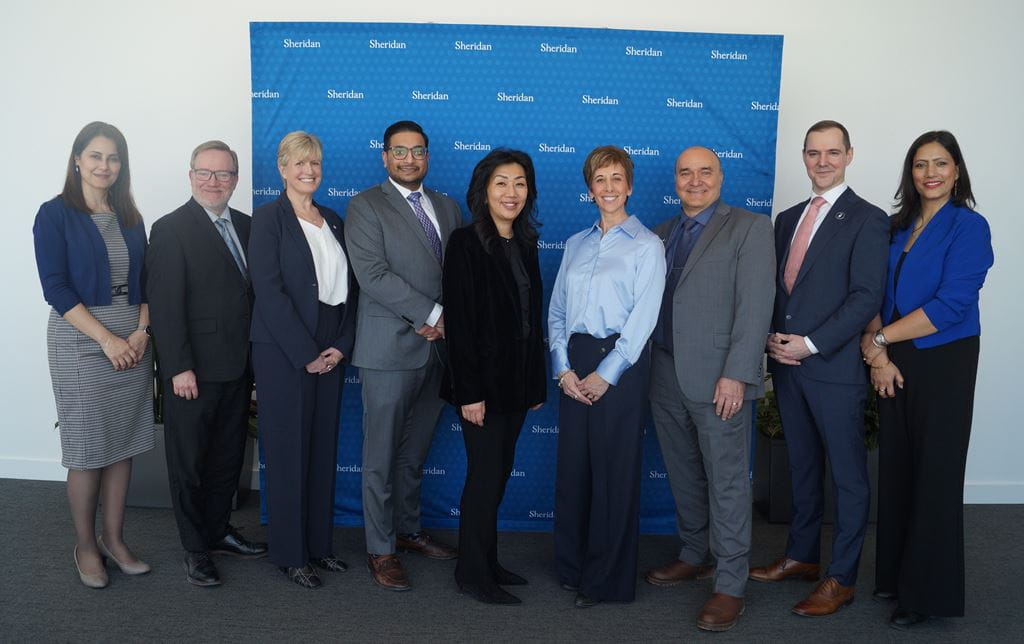 Nine people wearing business attire stand as a group in front of a blue Sheridan backdrop.