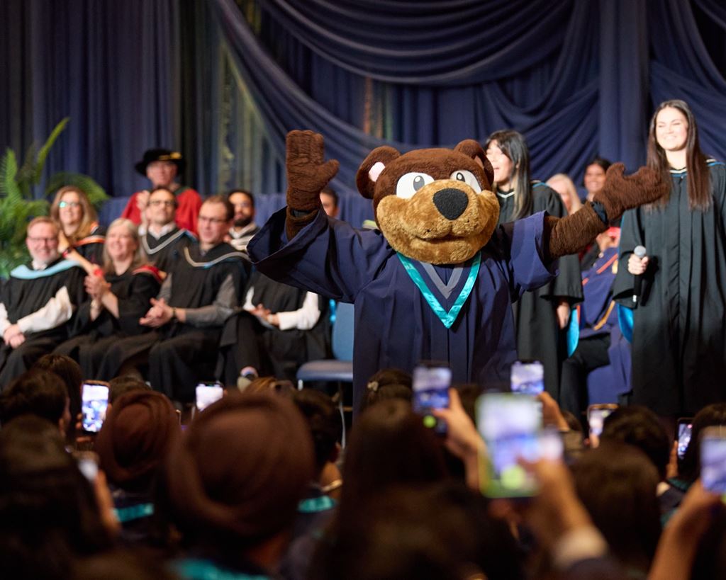 People stand on stage with Sheridan's mascot, Bruno, during a Convocation ceremony