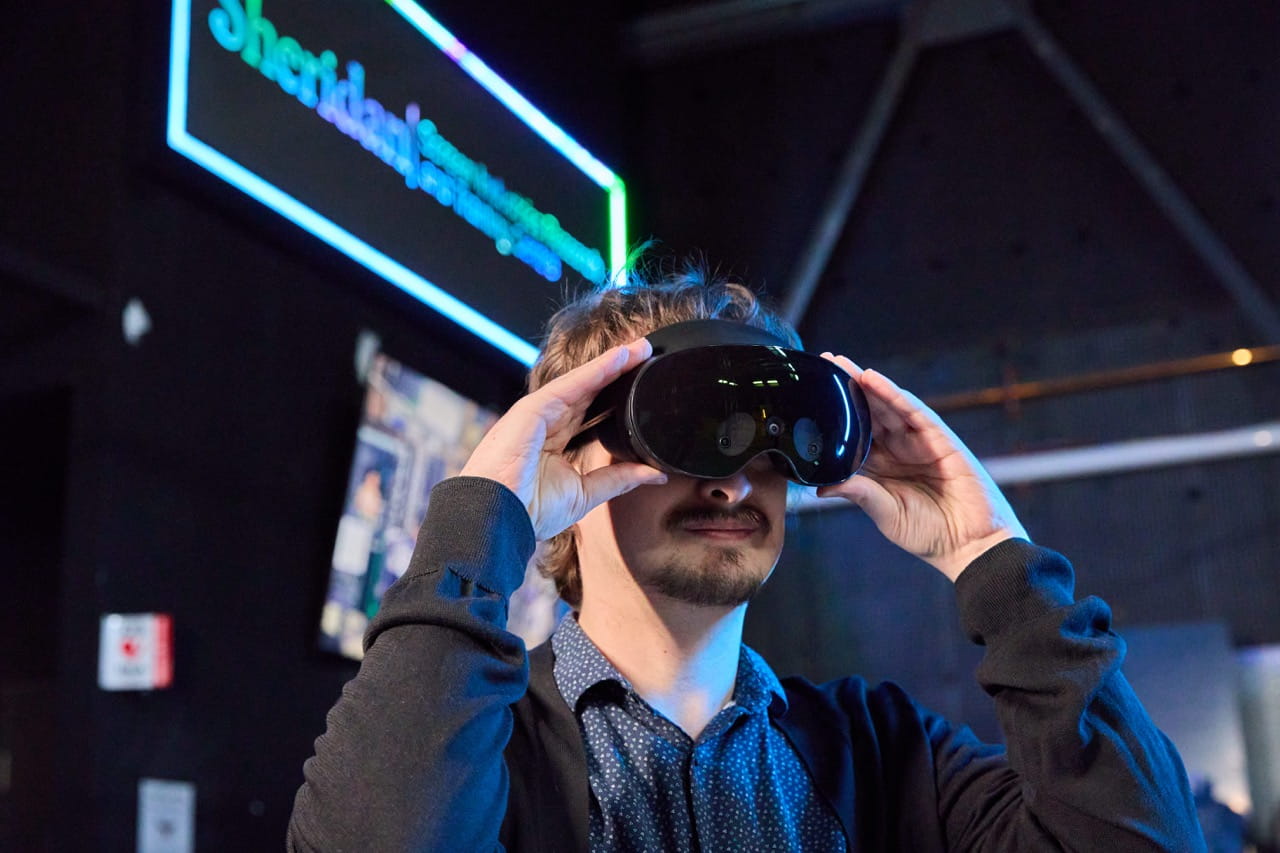 A person adjusts a black virtual reality headset while standing in a dimly lit technology exhibition space. Blue and green neon lighting illuminates the scene, with a glowing Sheridan-branded sign and display screens visible in the blurred background. The image highlights an immersive VR experience in a modern, innovation-focused setting.