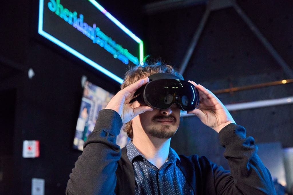 A person adjusts a black virtual reality headset while standing in a dimly lit technology exhibition space. Blue and green neon lighting illuminates the scene, with a glowing Sheridan-branded sign and display screens visible in the blurred background. The image highlights an immersive VR experience in a modern, innovation-focused setting.