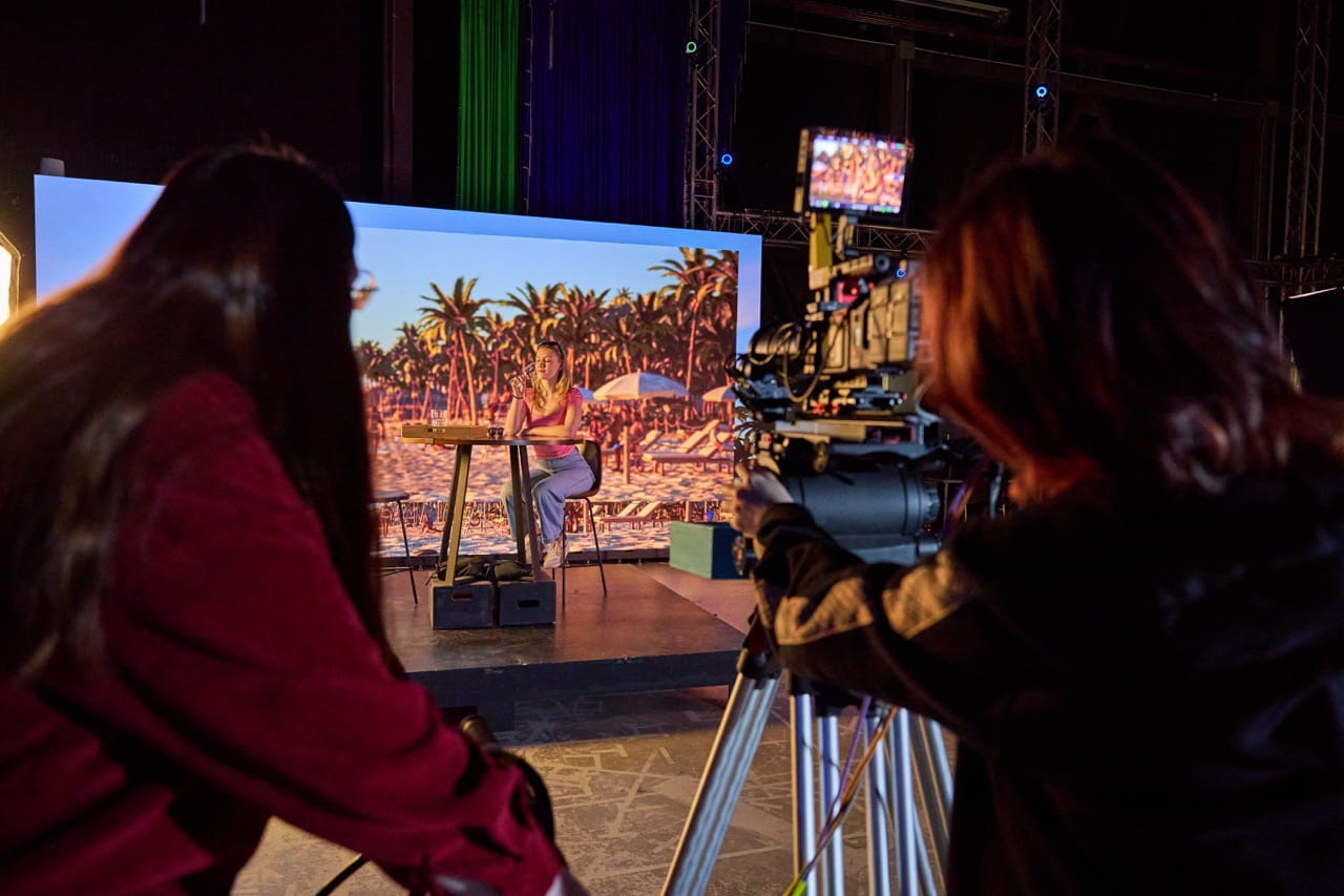 A camera operator films a person seated at a table on a studio set. Behind the subject, a large digital screen displays a tropical beach scene with palm trees and lounge chairs, demonstrating a virtual production environment. Production equipment, lighting structures, and crew members are visible in the foreground and background.