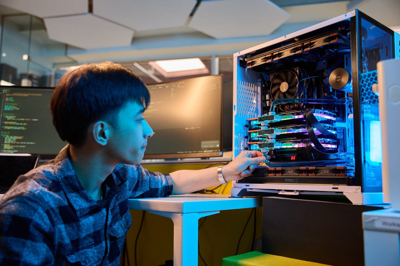 A person works beside an open desktop computer equipped with multiple graphics processing units (GPUs) illuminated by blue lighting. Several monitors displaying computer code are visible in the background, highlighting a computing environment used for artificial intelligence, machine learning, or advanced data processing.