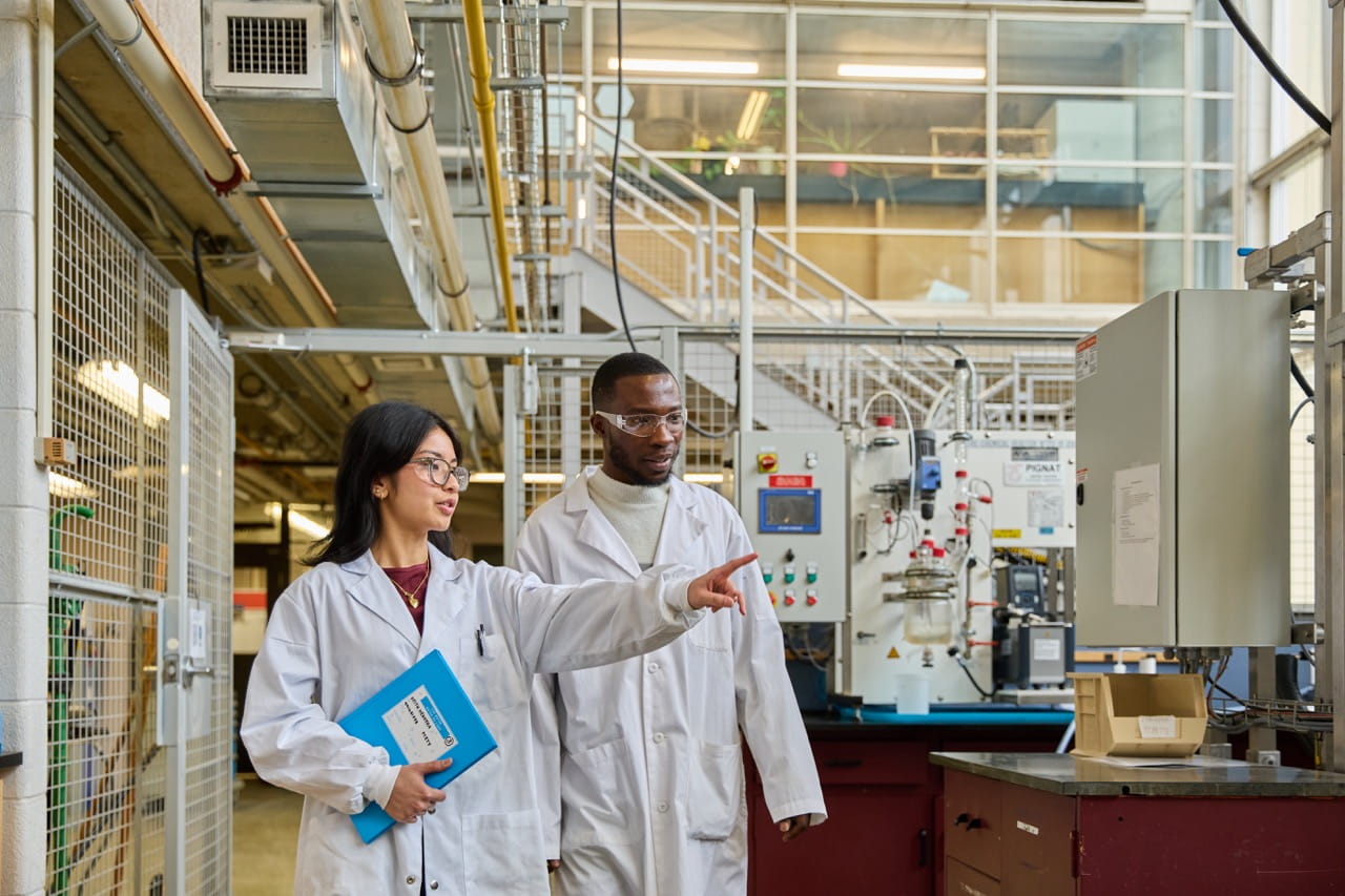 Two people wearing lab coats walk through an industrial research laboratory. One person holds a blue folder and points toward laboratory equipment and control systems. The facility contains machinery, piping, workstations, and safety infrastructure in a bright, modern research setting.