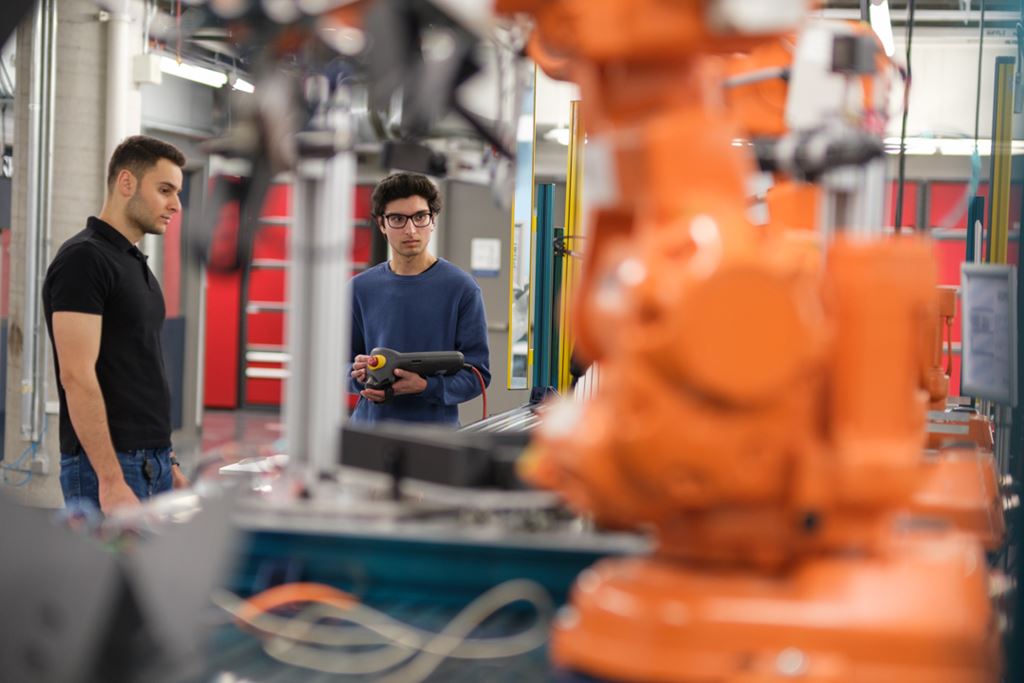 A large orange mechanical arm is pictured in Sheridan's Centre for Intelligent Manufacturing, with two staff members standing in the background