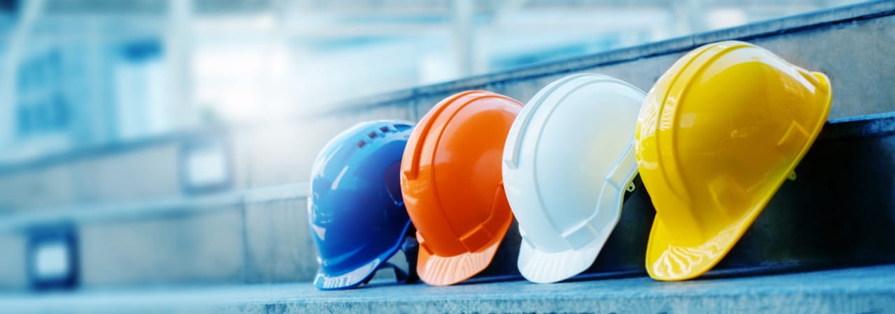 Blue, orange, white and yellow hardhats are leaned up against a metal step, representing diversity in construction