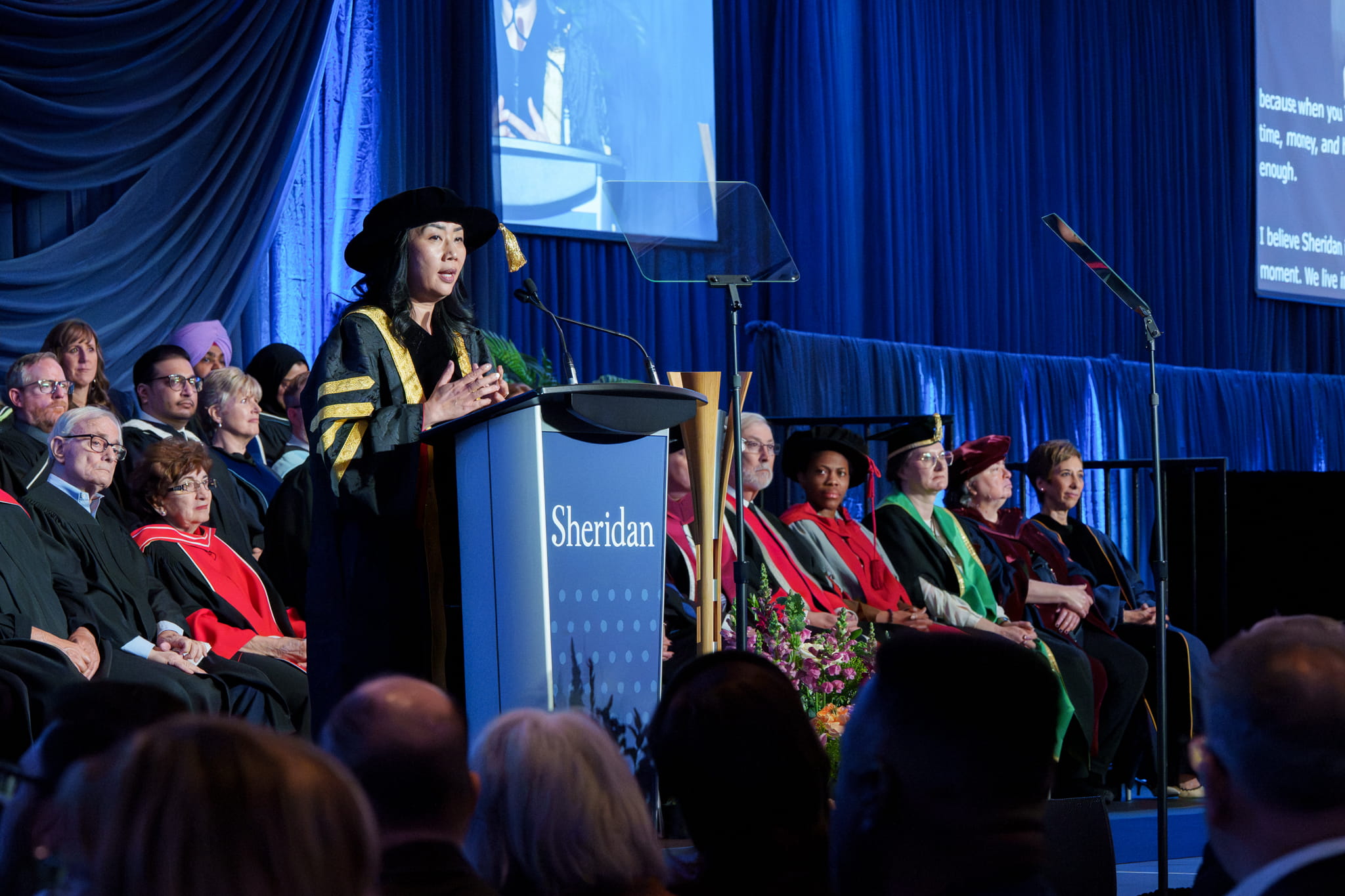 Sheridan President and Vice Chancellor Dr. Cindy Gouveia stands on stage delivering a speech from behind a Sheridan podium
