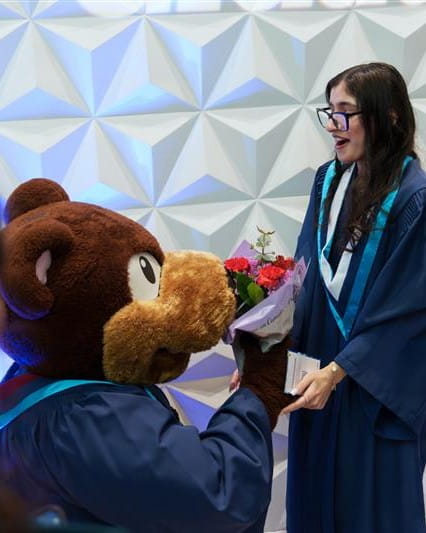 Bruno the bear mascot kneels and gives a graduating student a bouquet of flowers.